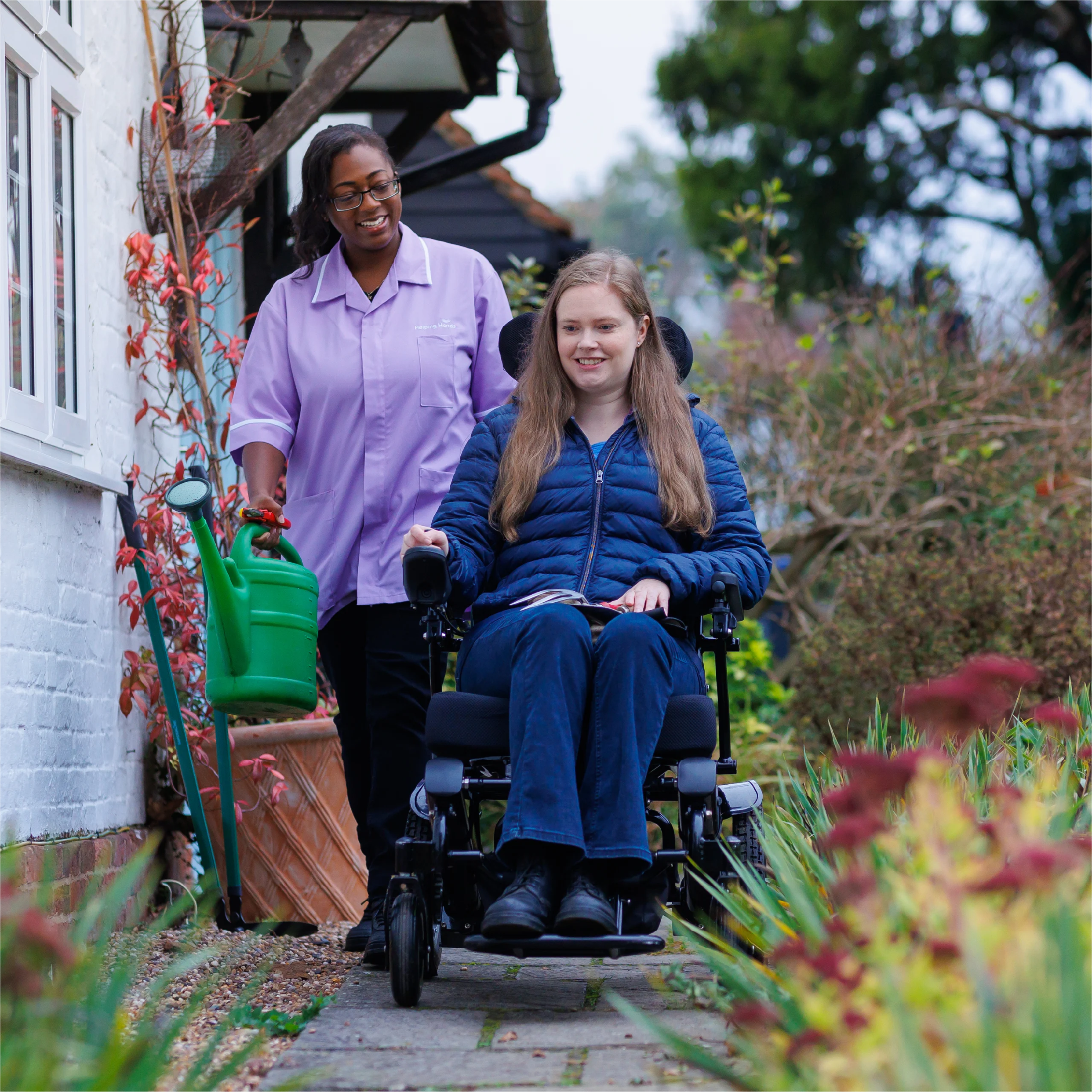 Support worker walking beside a young woman in a powered wheelchair along the garden path outside her home